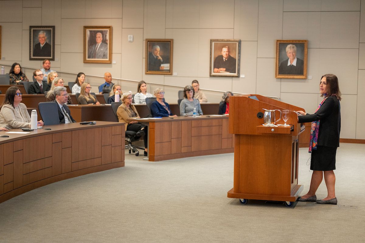 Woman speaking at a wooden podium to seated audience in a council chamber with portraits.