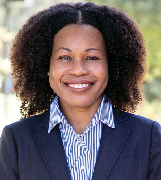 Stacy-Ann Elvy wearing a suit jacket and blue button-down. She is smiling brightly at the camera.