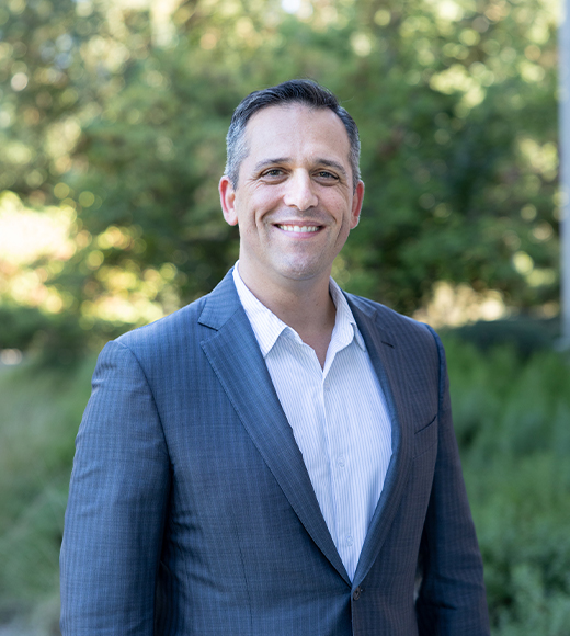 King Hall Alumni Board President Eric Toscano wearing a white button-down and grey suit jacket. He is smiling at the camera in front of green trees.