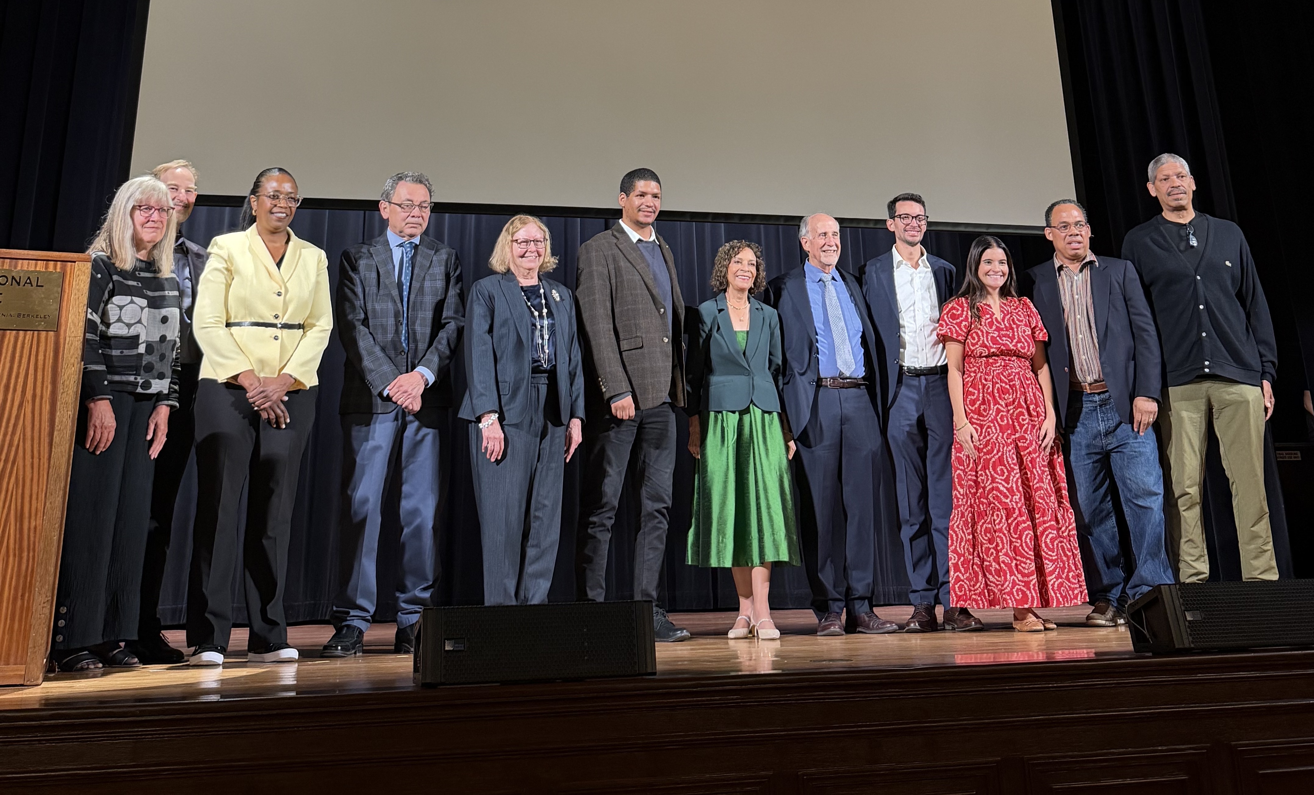 Group photo on stage of 11 smiling people in varied formal attire against plain backdrop