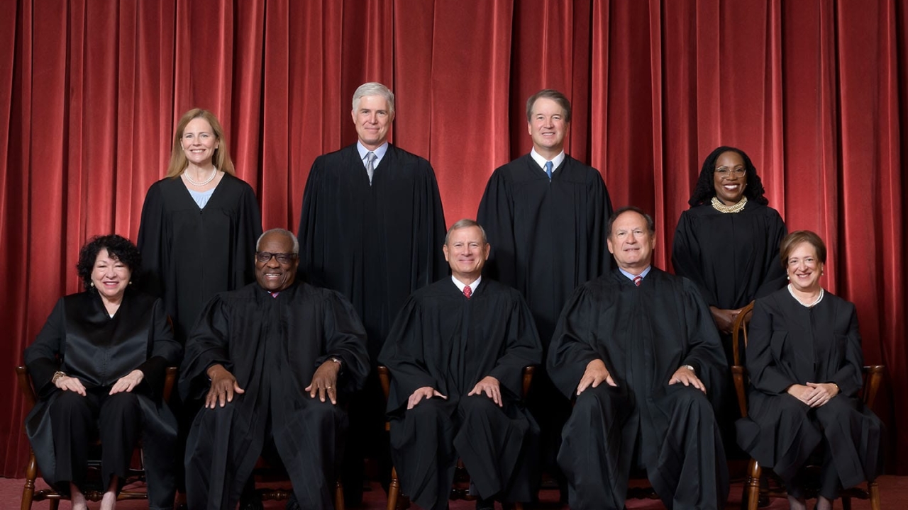 Nine judges in black robes posed in two rows before red curtains.