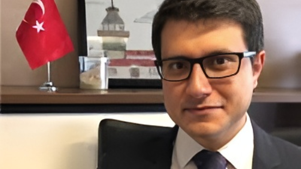A man in a suit with glasses sits in an office, a Turkish flag in the background.