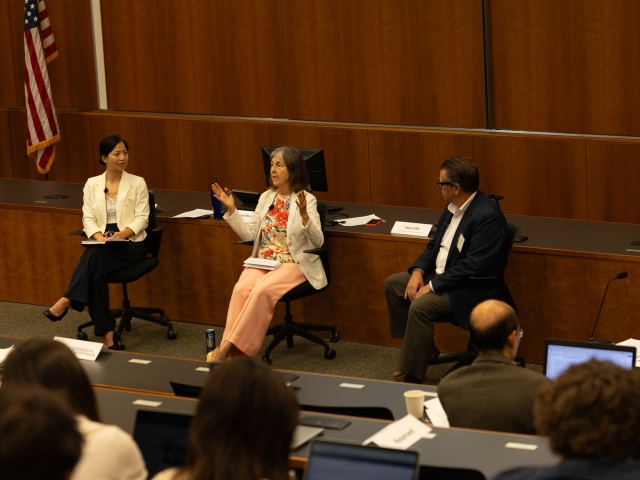 Three panelists speaking in a wood-paneled lecture hall with an American flag in the background.