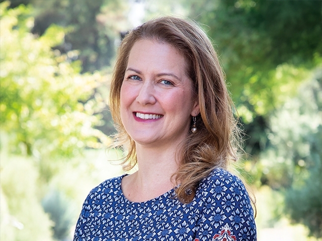 Senior Associate Dean Shestowsky wearing a blue blouse and smiling at the camera with green trees in the background.