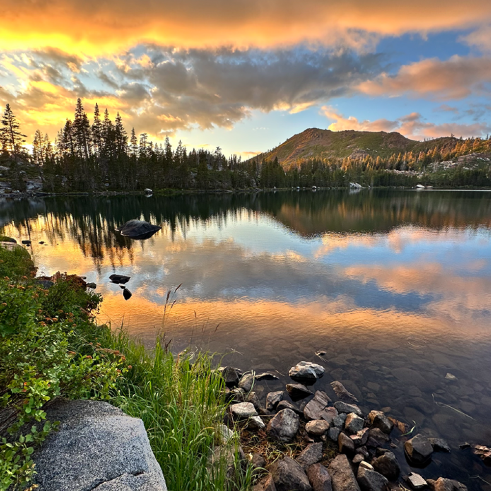 A serene lake at sunset reflecting colorful clouds and surrounding trees.