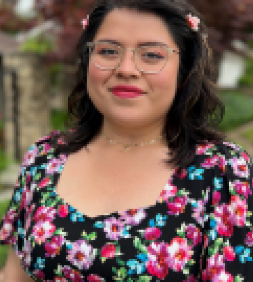 A woman wearing glasses and a colorful blouse with flowers. She is smiling at the camera.