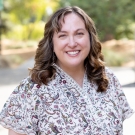 A smiling woman with curly brown hair wearing patterned blouse outdoors.